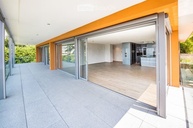 Modern balcony with large sliding glass doors leading to a kitchen area, featuring a light-colored tiled floor and a sleek, contemporary design.