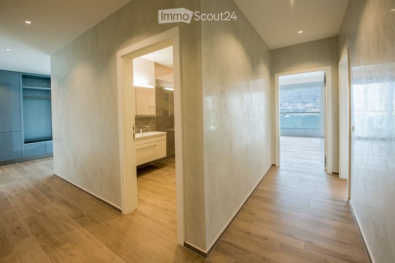 Modern hallway with wooden flooring, recessed lighting, and open doorways leading to a bathroom and a balcony area with a scenic view.