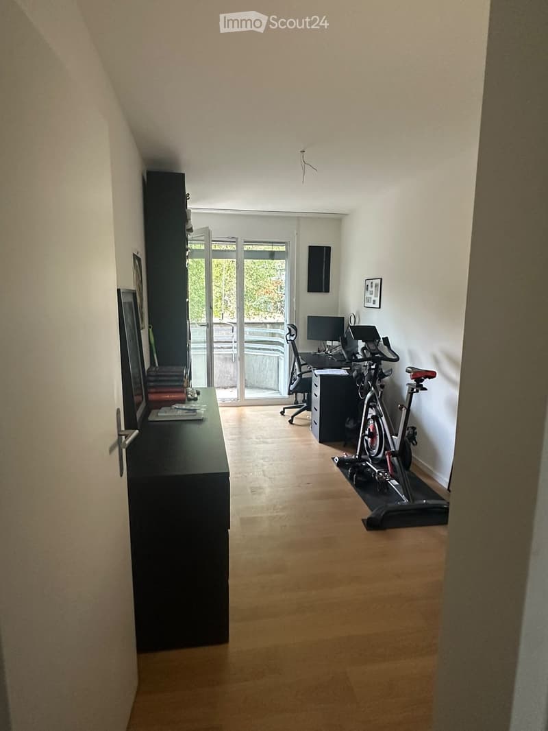 A modern, well-lit room with wooden flooring, a desk with a chair, an exercise bike, and a large window with a view of the garden.