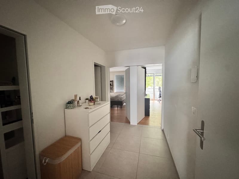 A narrow hallway with tiled flooring, white walls, and a ceiling light. A small wooden bench and a white dresser with various items on top are visible on the left side. An open doorway leads to a bedroom with a bed and a chair, and another doorway leads t