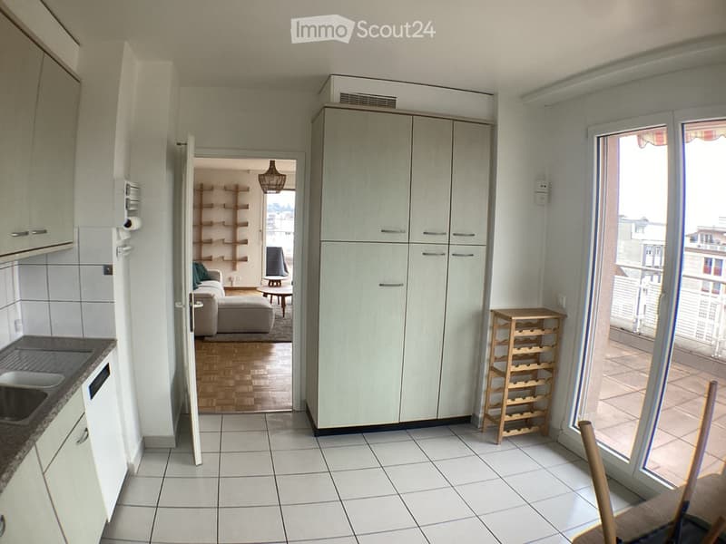 Modern kitchen with white tiled floor, white cabinets, sink, dishwasher, and a wine rack. An open doorway leads to a living area with a sofa, ottoman, coffee table, and a shelf unit.