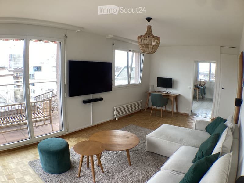 Modern living room with a grey sofa, wooden coffee tables, teal stool, and a large TV. Features a balcony with a view, a hanging wicker lamp, and a small desk area with a computer.
