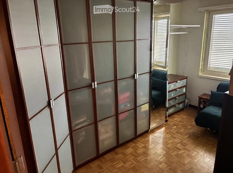 A furnished room with wooden flooring, a large sliding wardrobe, a sofa, a small table, and a drawer unit.
