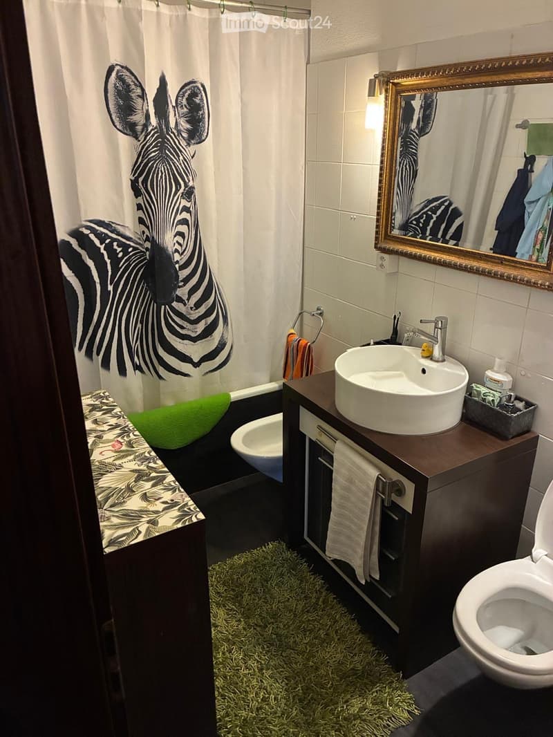 Bathroom with a zebra-patterned shower curtain, a sink with a modern faucet, a toilet, a mirror with a decorative frame, a towel, and a small bench with a green cushion.