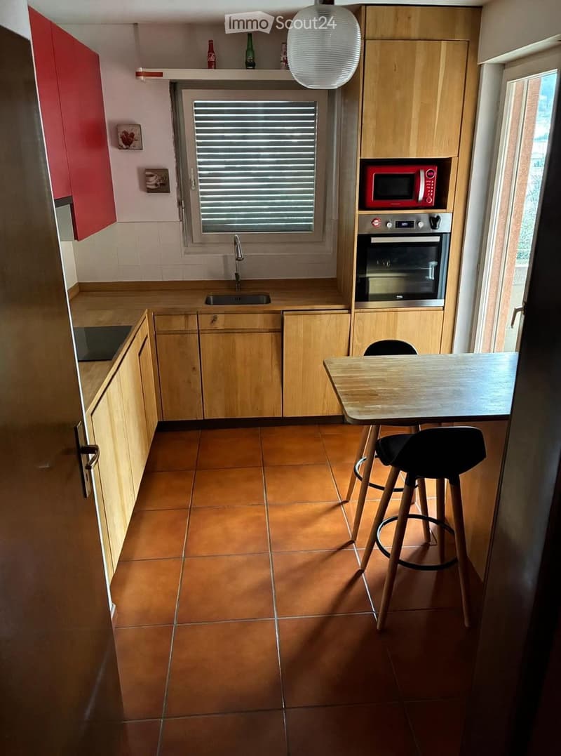 Modern kitchen with wooden cabinets, a red microwave, a built-in oven, a sink with a faucet, a small table with two stools, and a window with blinds.