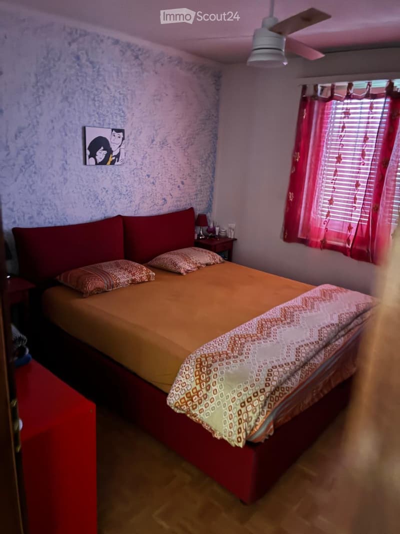 Cozy bedroom with a single bed, red headboard, patterned bedspread, nightstand with lamp, and a picture frame on the wall.