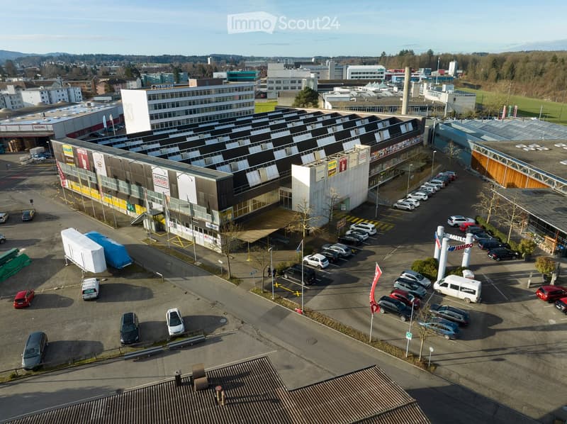 Aerial view of a commercial building complex with multiple storefronts, parking lot, and surrounding urban area.