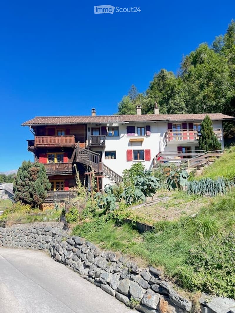 A multi-story house with a red-tiled roof, white walls, and red shutters. Features include balconies, a stone retaining wall, and a landscaped garden area.