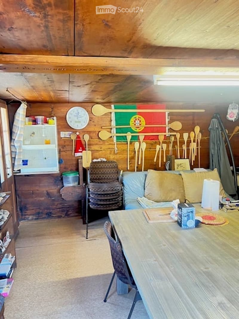 A cozy, rustic living space with wooden walls and ceiling, featuring a sofa, a wooden table with chairs, a small fridge, and decorative spoons on the wall.