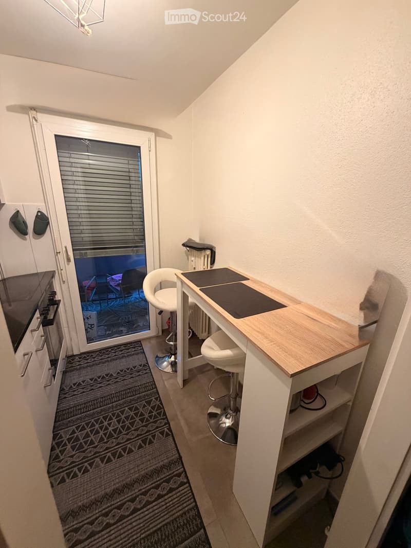 A small, modern room with a white stool, a wooden table, a radiator, an oven, and a patterned rug.