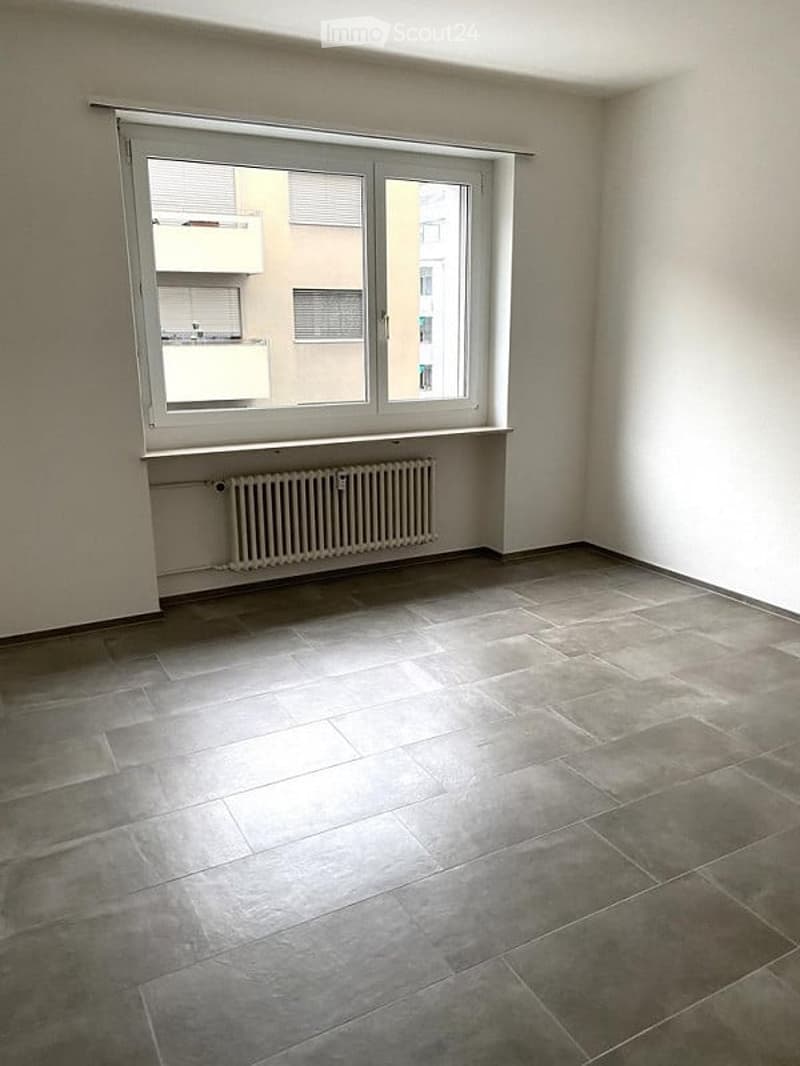 Empty room with tiled floor, white walls, large window with blinds, radiator under the window, and a view of neighboring buildings.