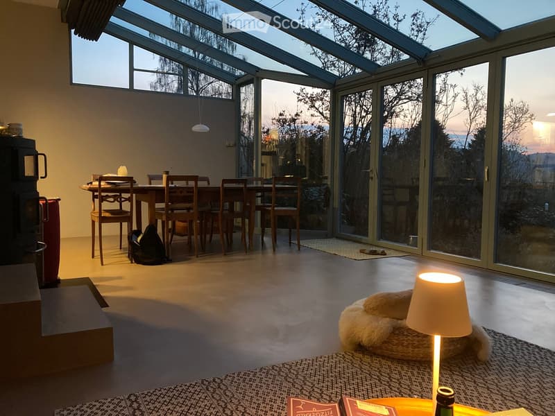 Bright, open room with large glass walls and a view, featuring a dining table with chairs, a dog bed, a lamp, and books on a table.
