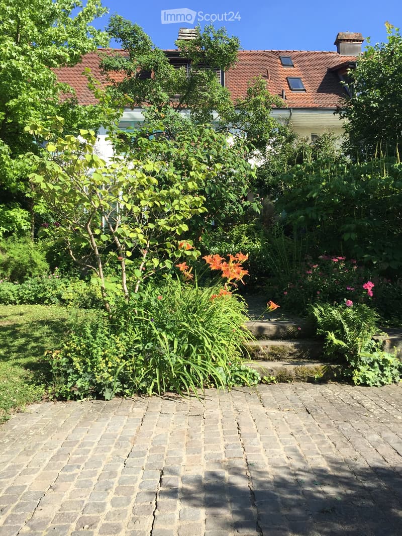 Garden with a stone-paved pathway, lush greenery, and colorful flowers, leading to a house with a red-tiled roof in the background.