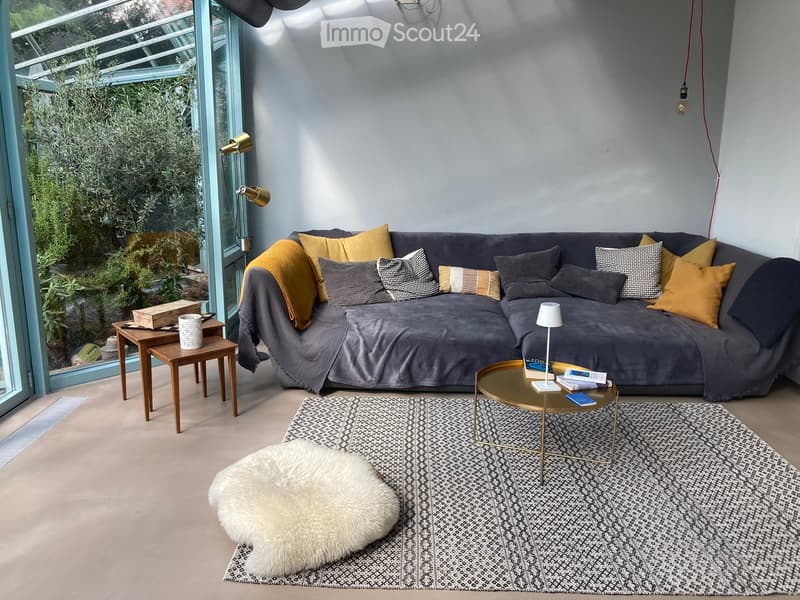 Modern living room with a grey sectional sofa, colorful throw pillows, a round gold coffee table, side tables, a white floor lamp, and a patterned rug.