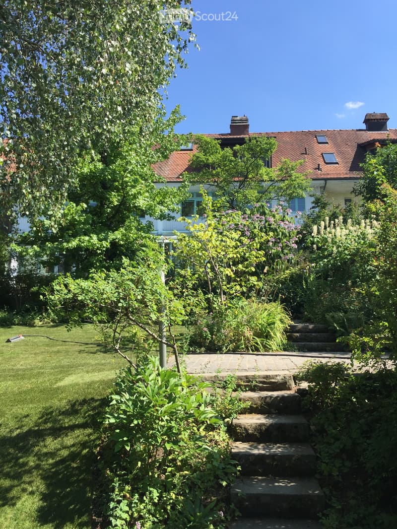 Garden with lush greenery, stone steps leading up to a house with a red-tiled roof and white walls.