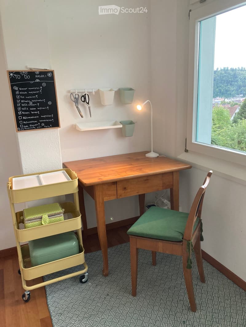 A small, well-lit room with a wooden desk, a green chair, a white lamp, a yellow cart with storage compartments, and a blackboard with a to-do list. The room has a large window with a scenic view and a patterned rug on the floor.