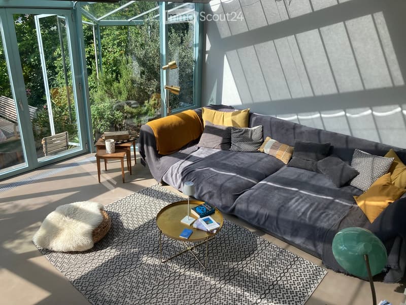 Modern living room with large windows, grey sectional sofa, colorful pillows, coffee table, side table, floor lamp, and books.