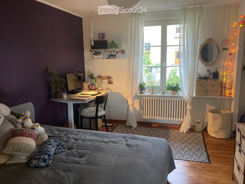 Cozy bedroom with a large bed, desk area, and window with plants. Features a purple accent wall, patterned rug, and decorative items on shelves.