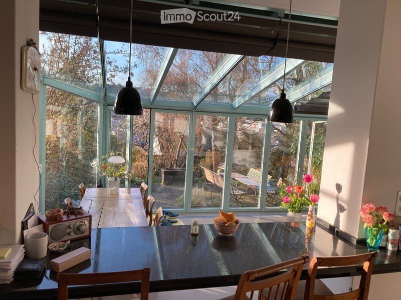 Bright, airy dining area with large glass walls and a skylight, featuring a black dining table with chairs, vases with flowers, and a bowl of fruit on the table.