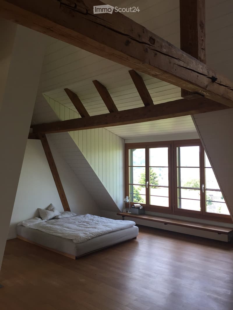 Bright attic bedroom with wooden beams, large windows, a bed, a nightstand with a lamp, and a wooden bench under the windows.
