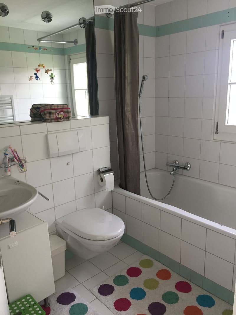 Bathroom with white tiles, a bathtub with a shower curtain, a toilet, a sink, a colorful rug, and a mirror above the sink.