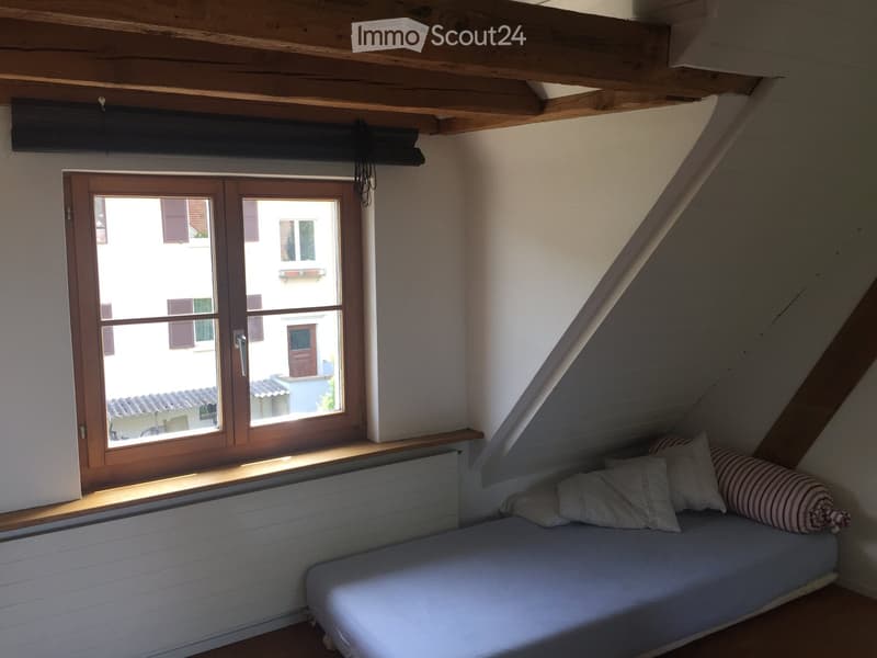 Cozy attic bedroom with a single bed, pillows, wooden window frame, and slanted wooden ceiling beams.