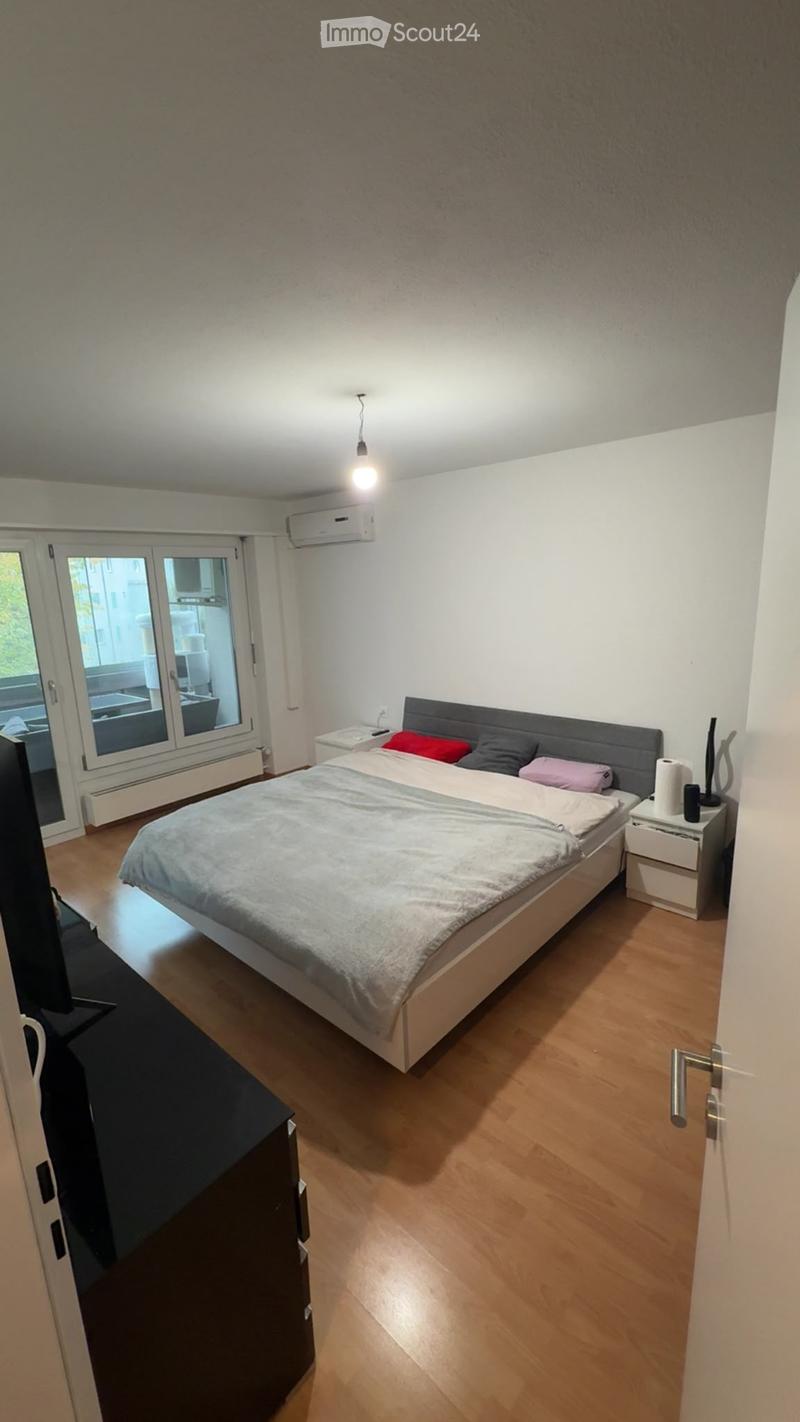 Modern bedroom with a large bed, nightstand, and a large window with a view of the balcony. The room features wooden flooring and white walls.