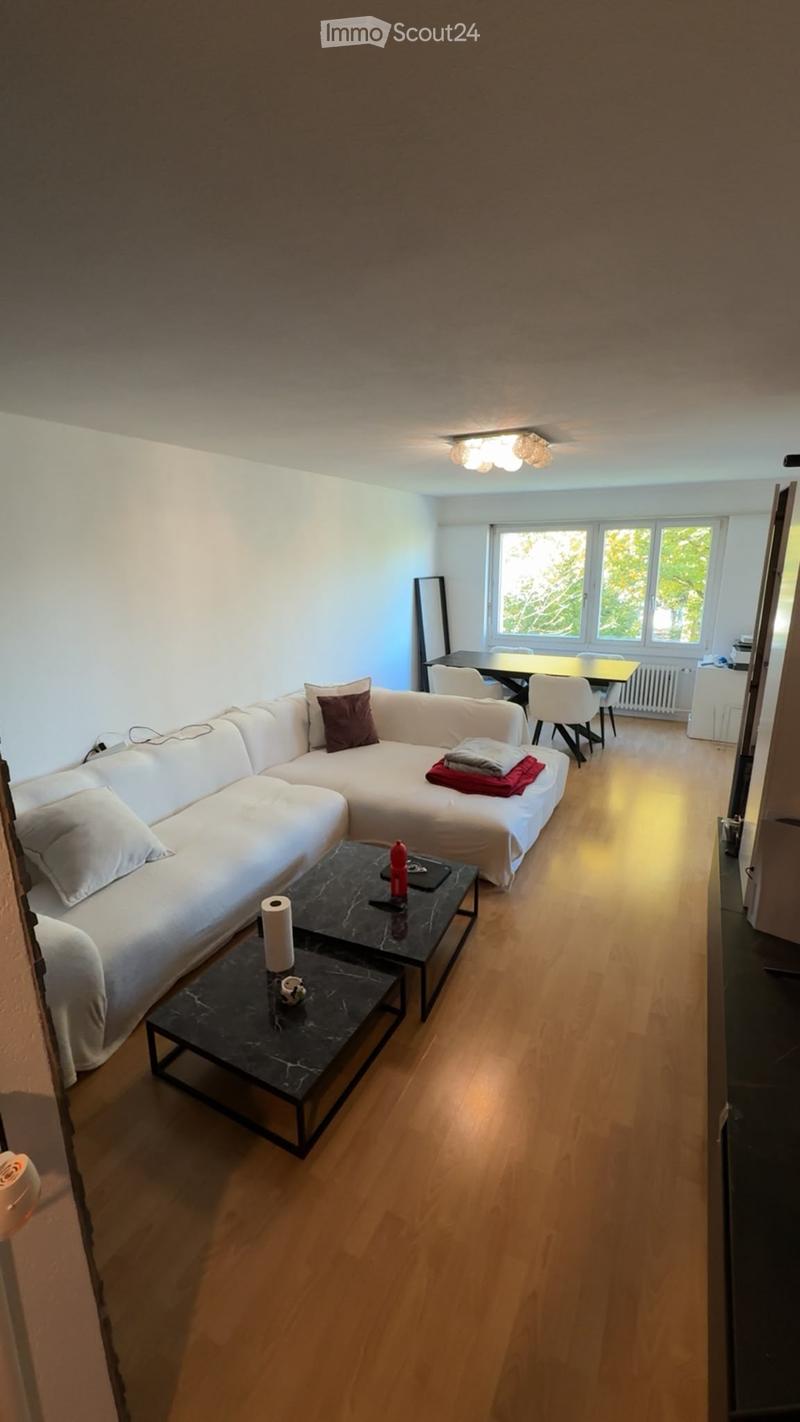 A modern living room with a white sectional sofa, a black coffee table, a dining area with chairs and a table, and large windows letting in natural light.