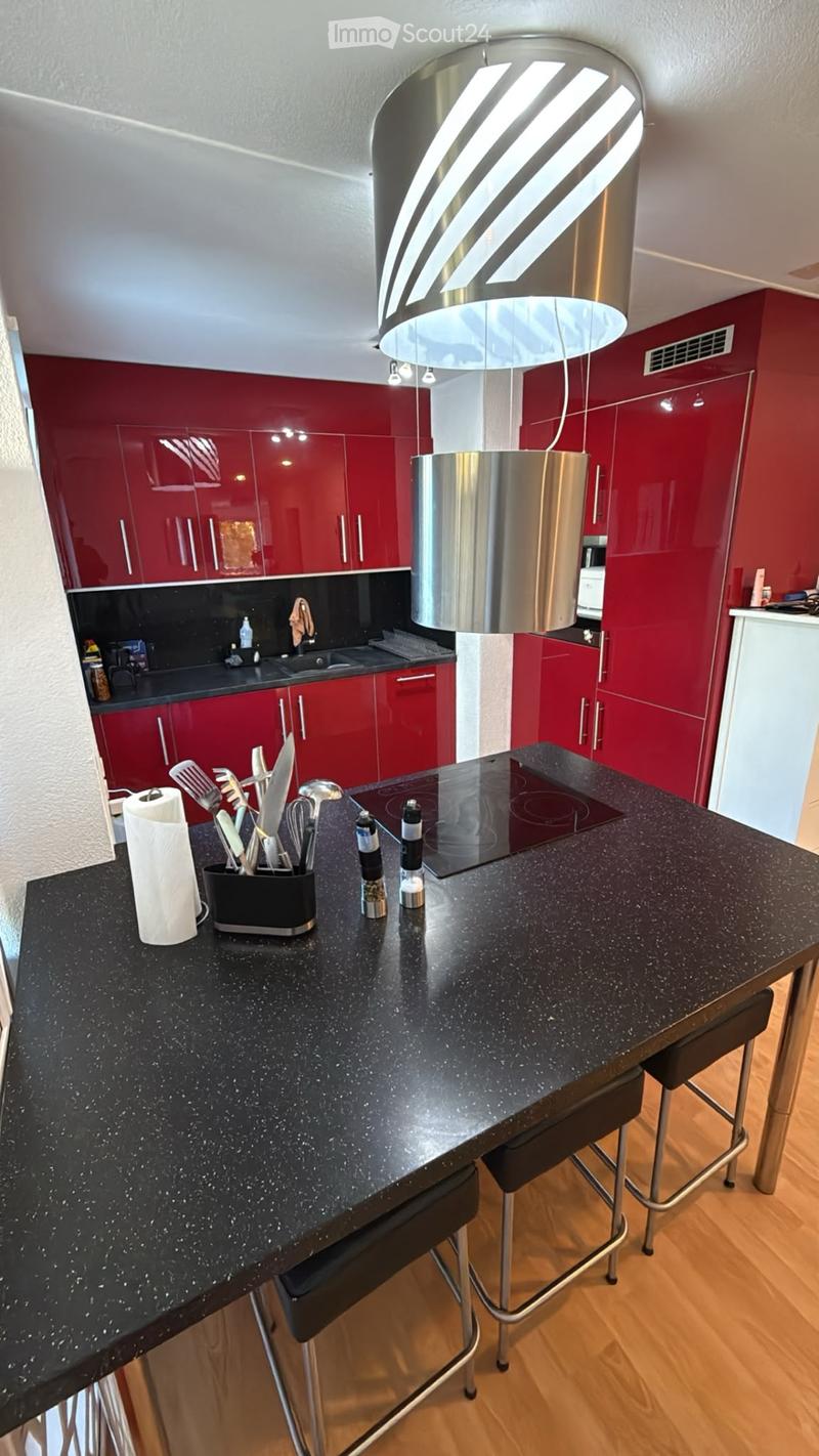 Modern kitchen with red cabinets, black countertop, induction cooktop, and a hanging pendant light. Kitchen utensils and a paper towel are on the counter.
