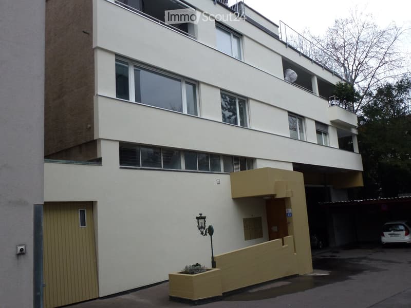 Modern multi-story building with large windows, balconies, and a small garden area in front.