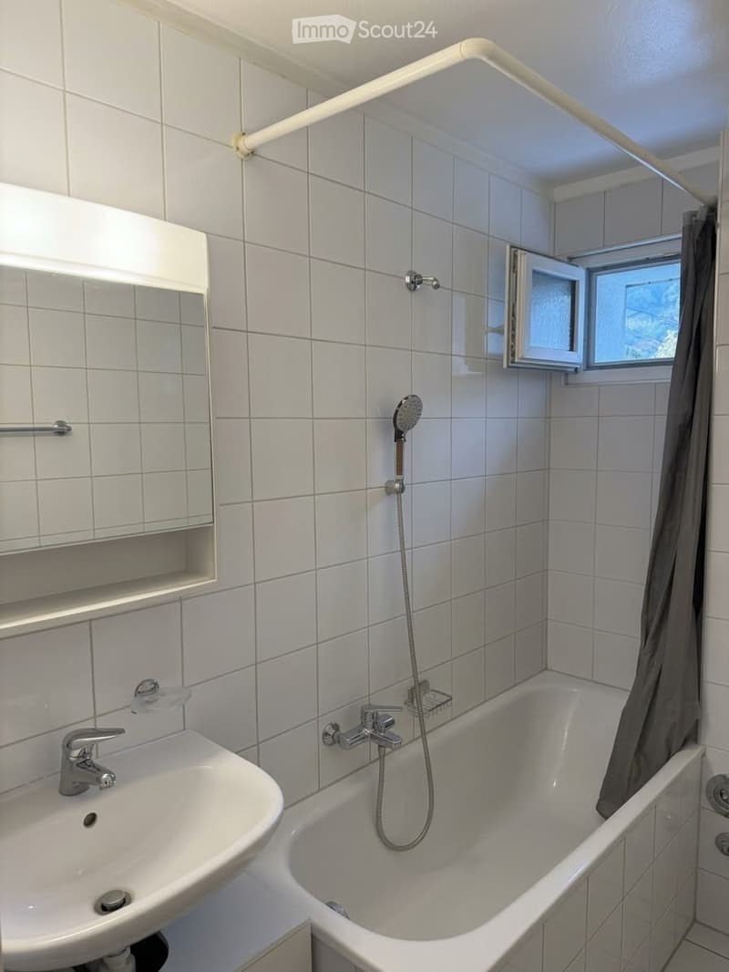 Modern bathroom with white tiles, a bathtub with a handheld shower, a sink with a mirror, and a window with a curtain.
