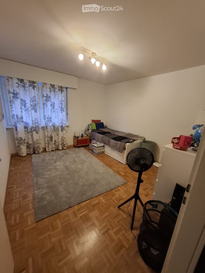 Cozy bedroom with wooden flooring, a bed with pillows and blankets, a nightstand, a fan, a large rug, and a window with floral curtains.