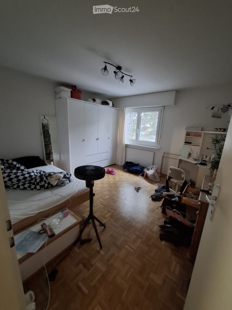 Bedroom with wooden floor, large window, bed with checkered blanket, wardrobe, fan, chair, desk, and mirror.