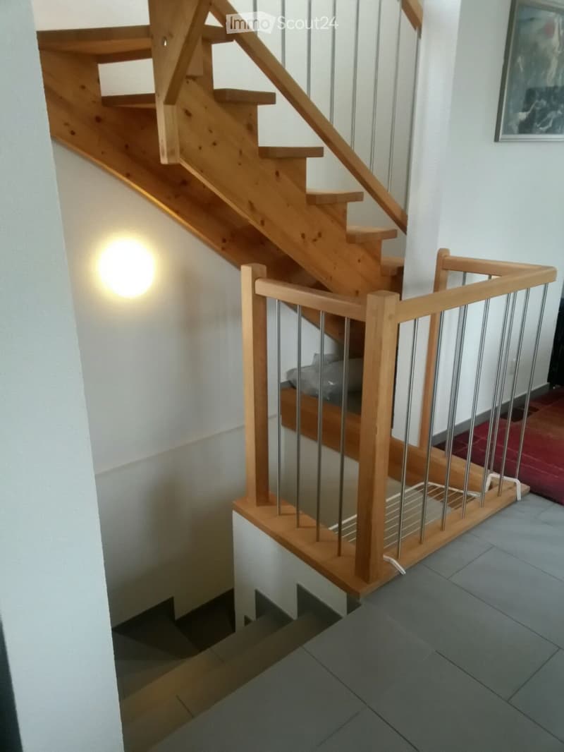 Wooden staircase with metal railing, modern lighting fixture on wall, grey tiled floor, white walls.