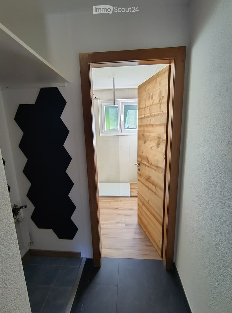 Modern hallway with grey tiled floor, white walls, wooden door frame, and a view into a room with wooden flooring and white walls.