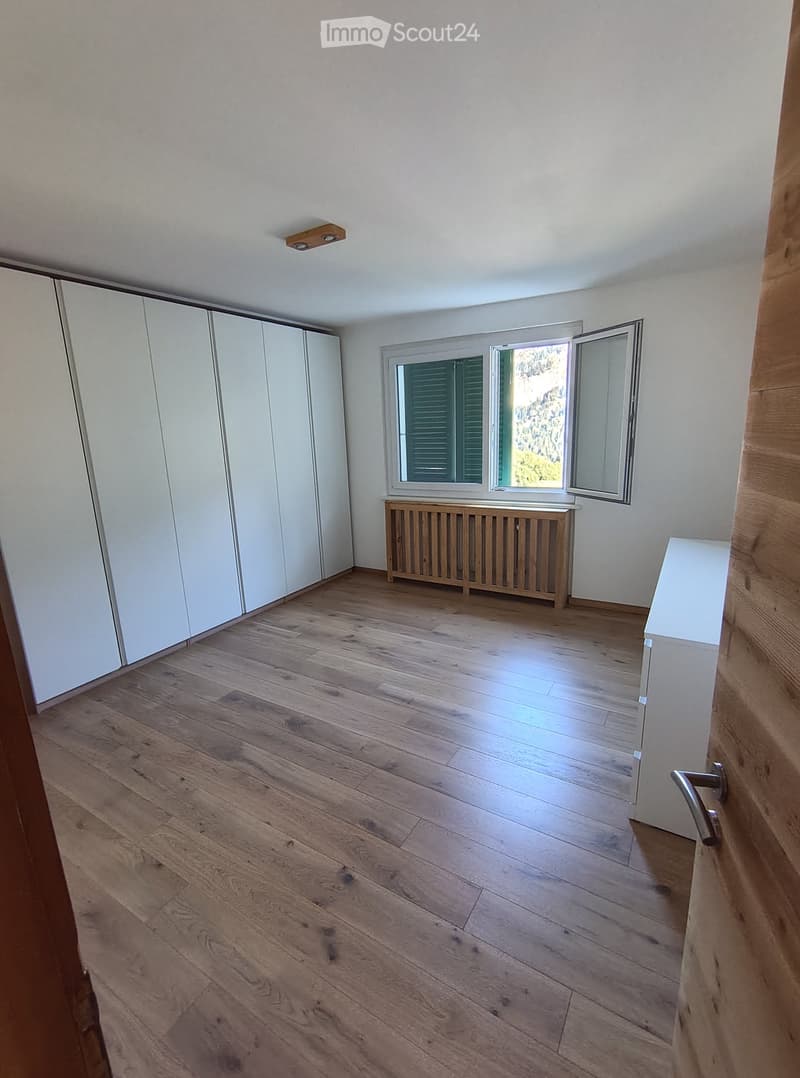 Empty room with wooden flooring, white walls, large window with shutters, radiator, and built-in white closet.