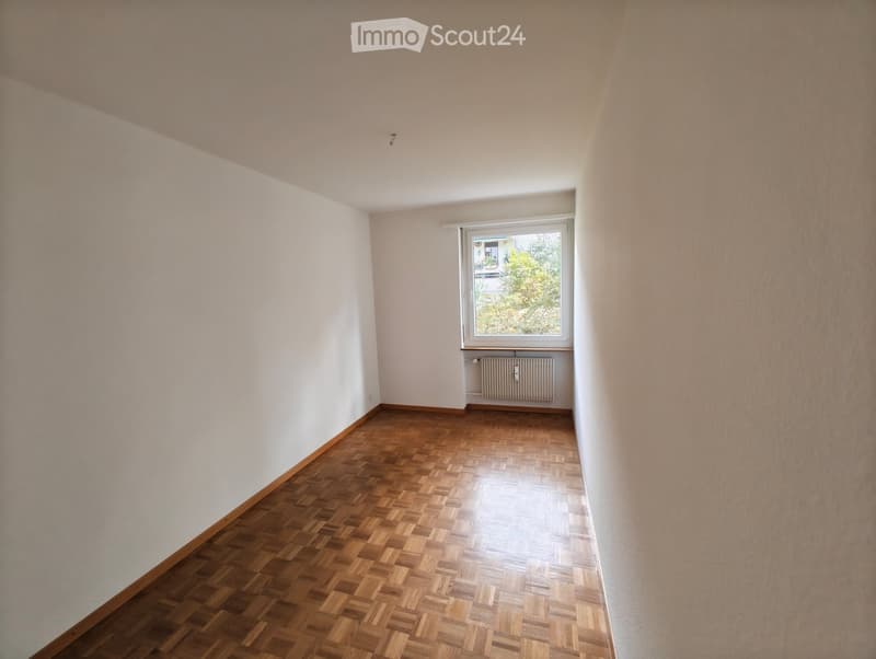 Empty room with wooden parquet flooring, white walls, and a single window with a radiator below it.
