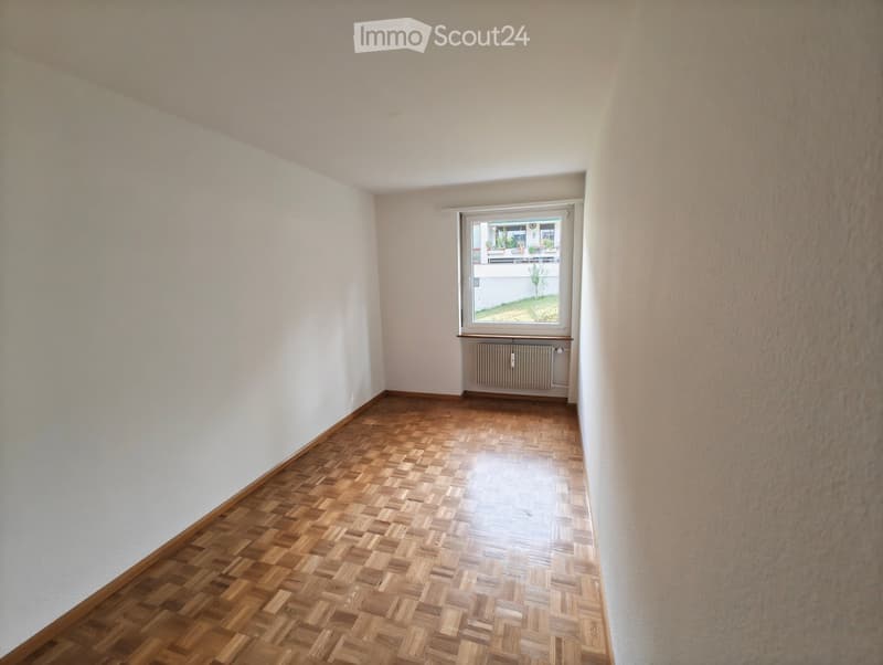 Empty room with wooden herringbone flooring, white walls, and a window with a radiator below it.