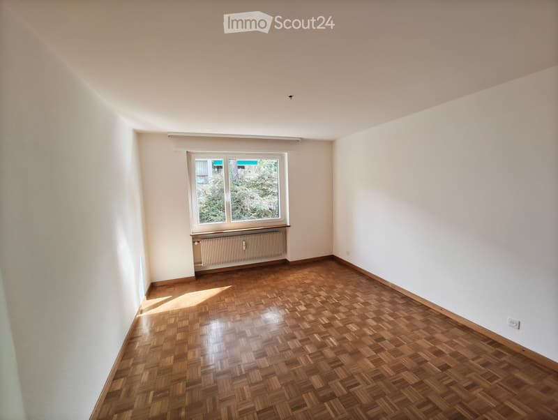Empty room with wooden parquet flooring, white walls, and a large window with a view of greenery. Radiator is visible below the window.