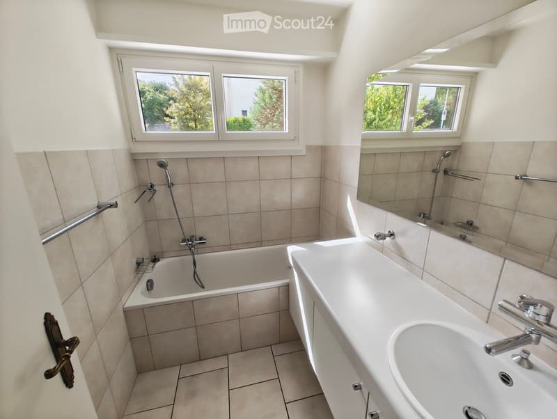 Modern bathroom with a bathtub, sink, mirror, and showerhead. Features tiled walls and a large window letting in natural light.