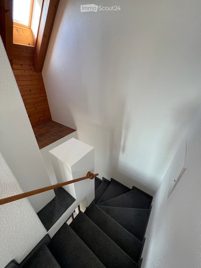 Modern staircase with dark grey carpeted steps, white walls, wooden handrail, and a small landing area with a window overlooking a wooden floor area.