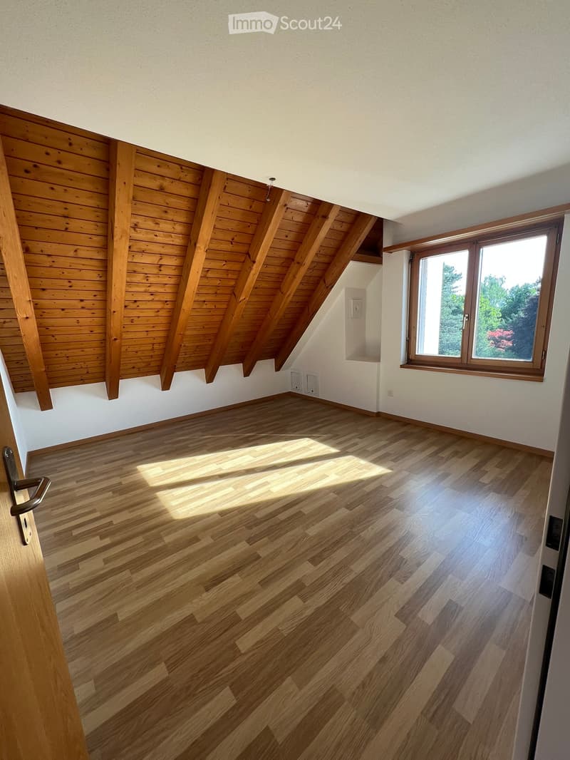 Empty attic room with wooden slanted ceiling, wooden floor, and large window letting in natural light.