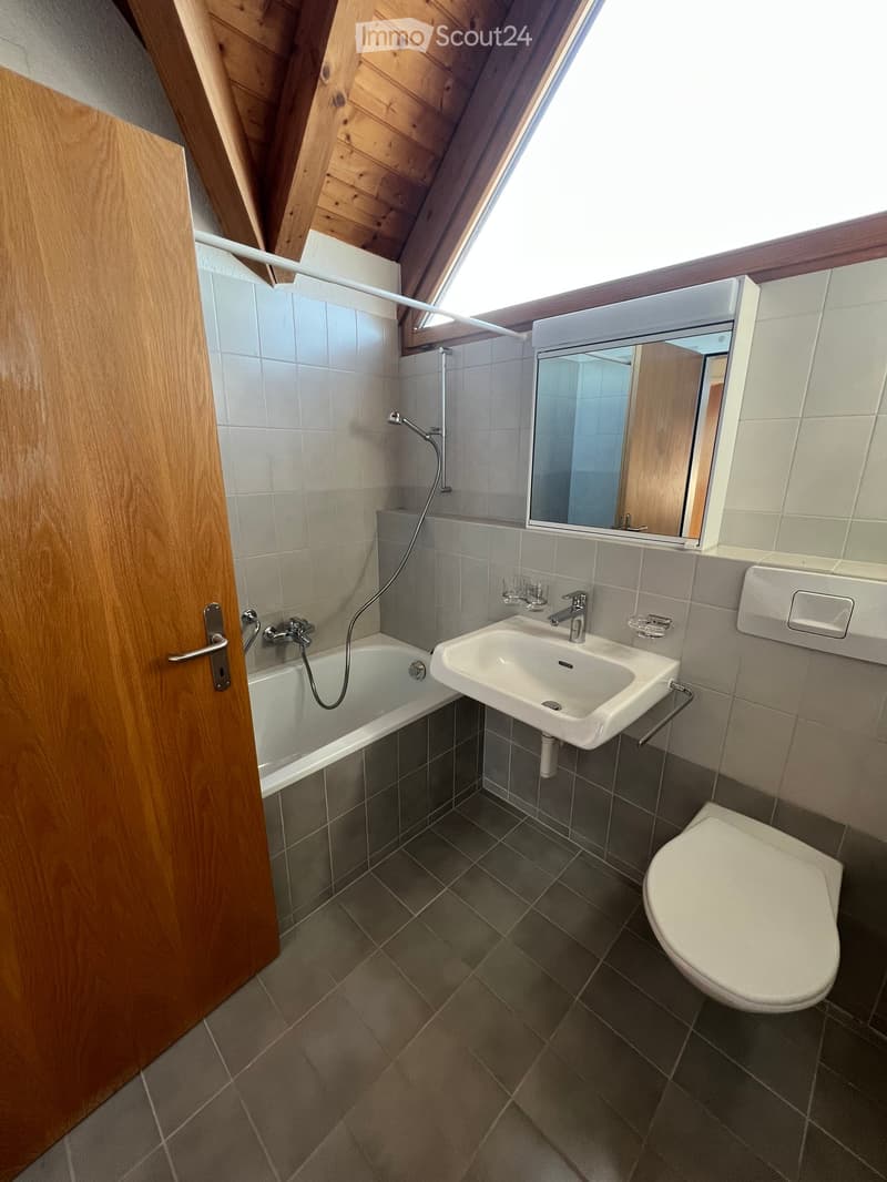 Modern bathroom with a bathtub, sink, toilet, and mirror. Features wooden ceiling beams and tiled walls.