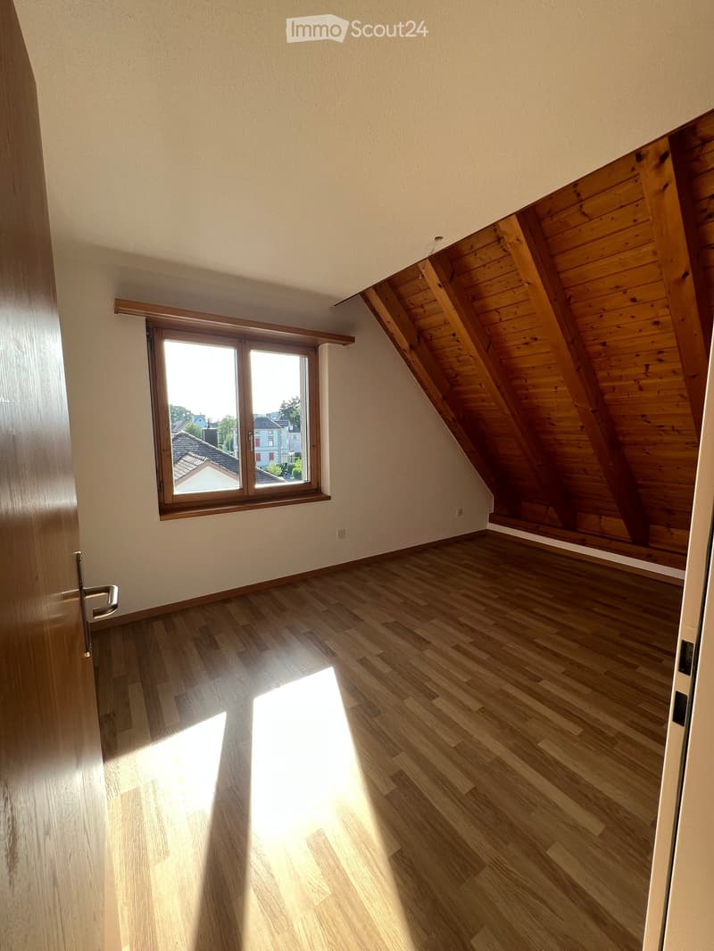 Empty attic room with wooden floors, slanted wooden ceiling, and large window offering a view of rooftops and trees.