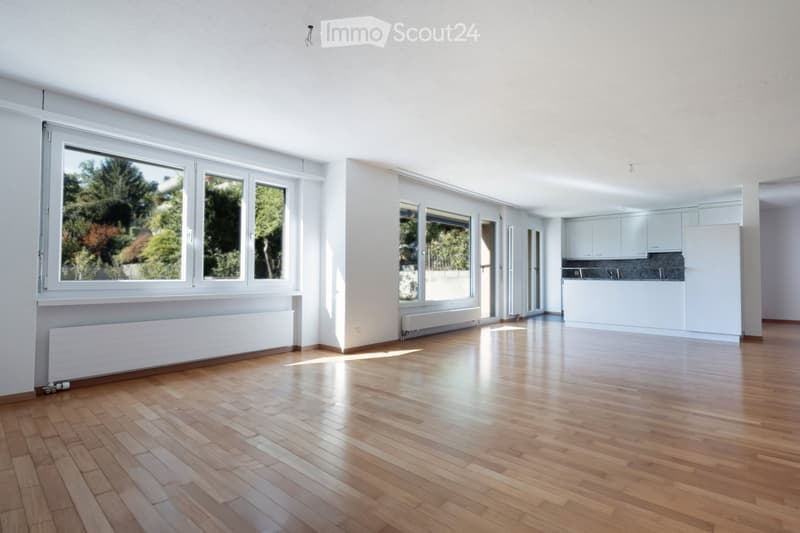 Bright, empty room with wooden flooring, large windows, and white walls. The space is clean and unfurnished, with natural light streaming in.