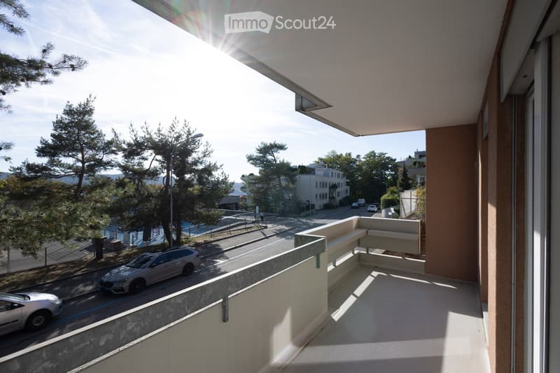 Balcony with modern seating area, overlooking a street with trees, parked cars, and distant buildings.