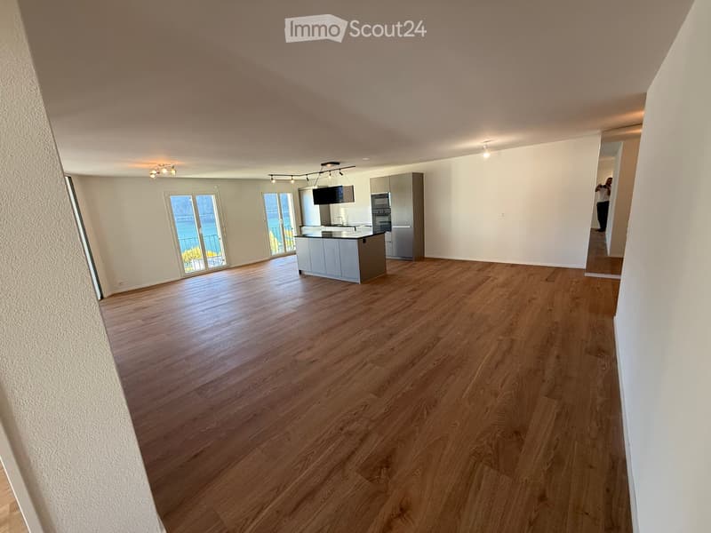 Open-plan living and kitchen area with wooden flooring, modern kitchen island, refrigerator, oven, and television. Large windows offer a view of the water.