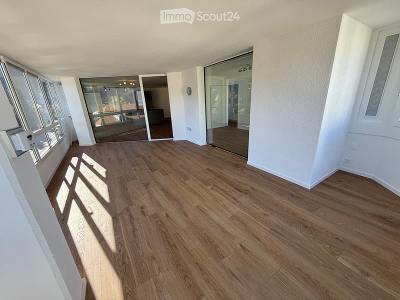 Empty room with wooden flooring, large windows, and glass sliding doors leading to another space.