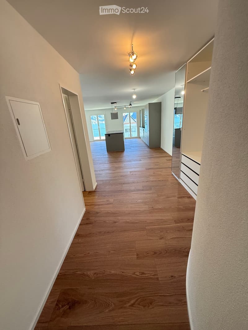 Empty hallway with wooden flooring, white walls, and built-in storage on one side. The hallway leads to a room with a kitchen area visible in the background.