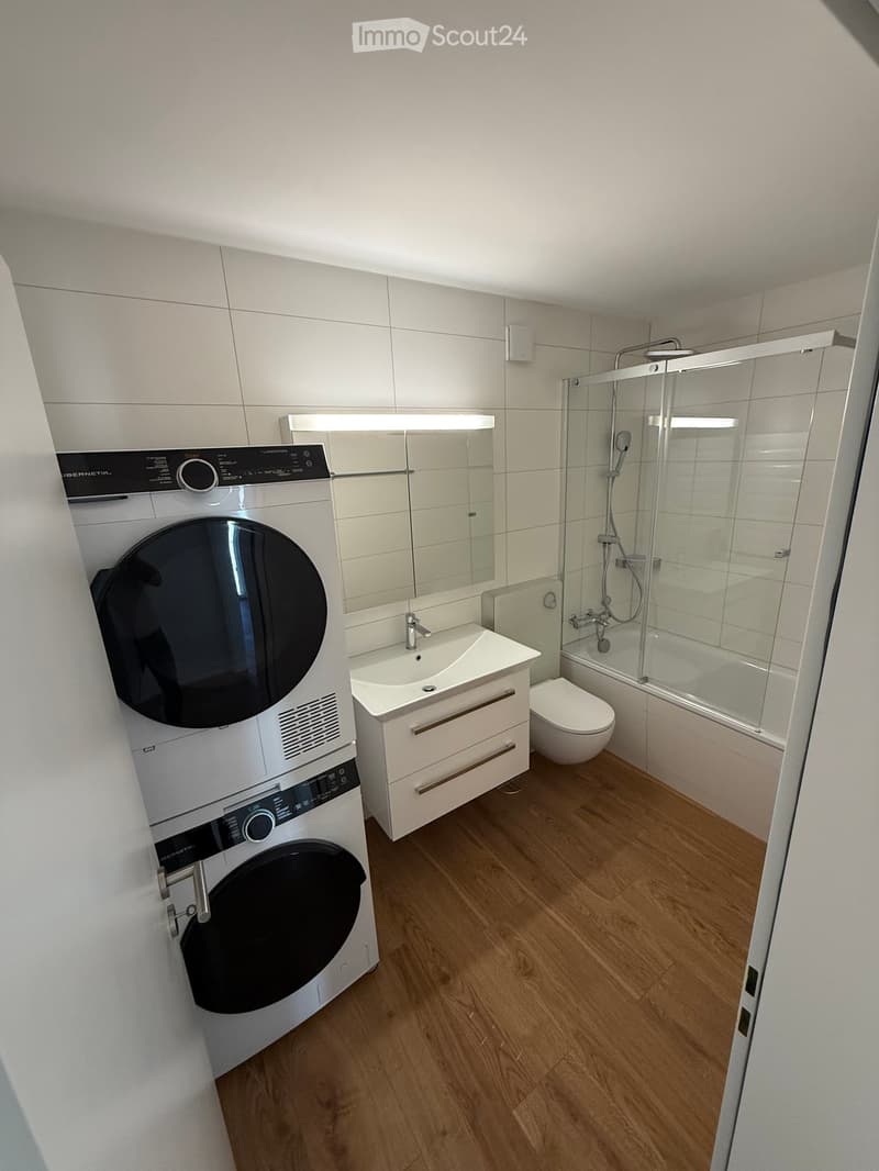 Modern bathroom with wooden flooring, white tiles, a washing machine, a sink with a mirror, a toilet, and a glass-enclosed shower.